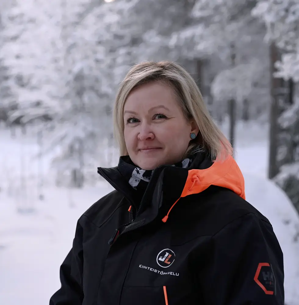 Nainen hymyilee lumisessa metsässä, yllään musta ja oranssi talvitakki kiinteistöpalvelu-logolla.